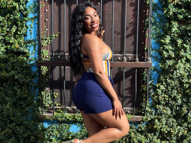 NatashaSparks with long black wavy hair, smiling, wearing a striped top and blue skirt, showing her curvy ass and small tits, standing in front of a wooden door.