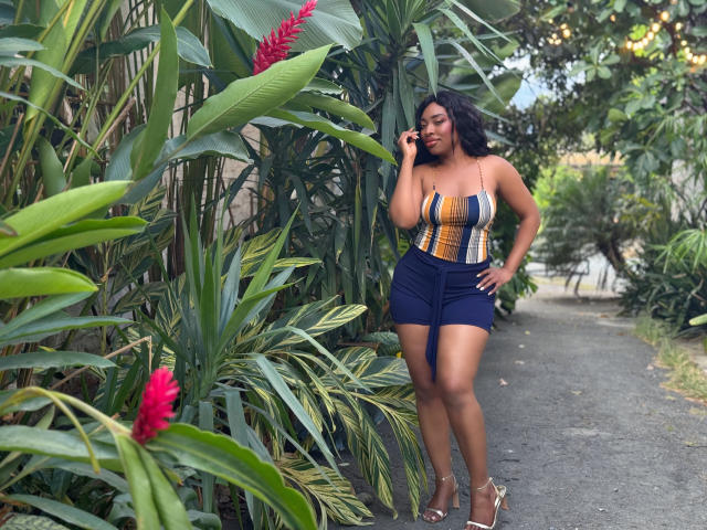 NatashaSparks with dark curly hair, talking on phone, wearing yellow-blue-striped top, blue high-waisted shorts, and gold heels, standing on path with lush green plants and red flowers.