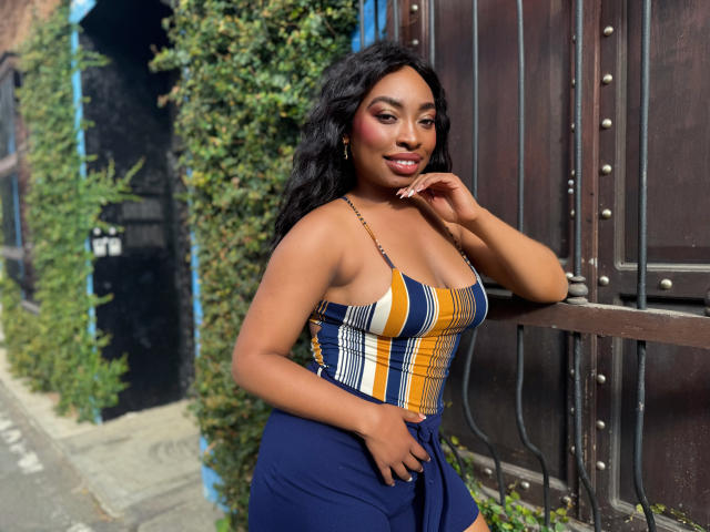 NatashaSparks with dark skin, curly black hair, and a cute smile, wearing a yellow, white, and blue striped top, showing her tits, and a blue skirt, leaning on a wall.
