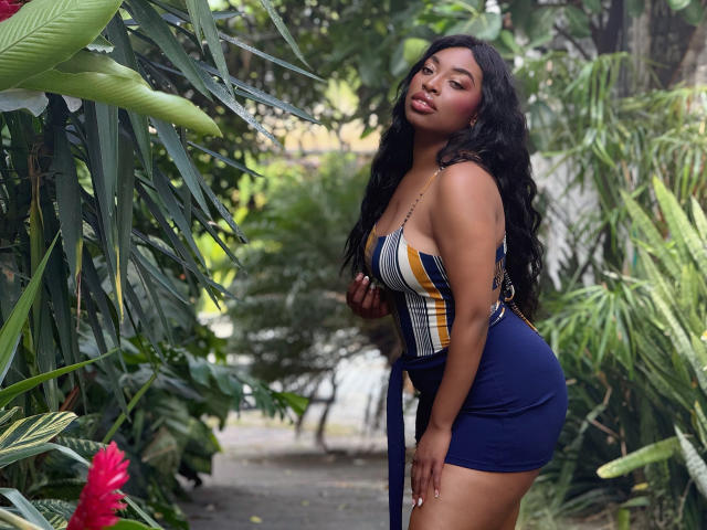 NatashaSparks with long black hair, wearing a blue and yellow plaid dress, showing her ass and tits, standing in a green, plant-filled area, looking at the camera.