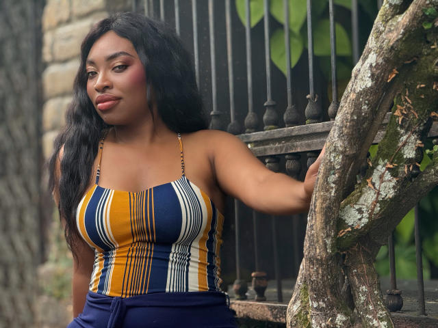 NatashaSparks with long black hair, wearing a striped yellow, blue, and white top, showing her big tits, leaning on a tree with a metal fence behind her.