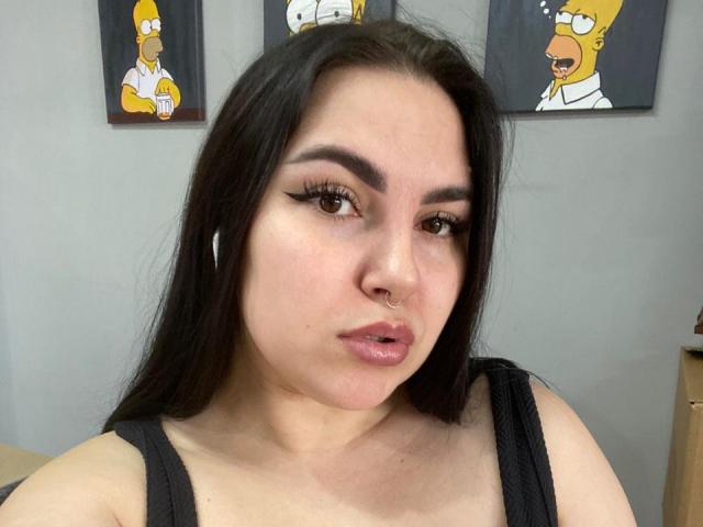 CisEva has long black hair, dark eyebrows, winged eyeliner, a nose ring, and glossy pink lips. She wears a black tank top, showing her boobs.