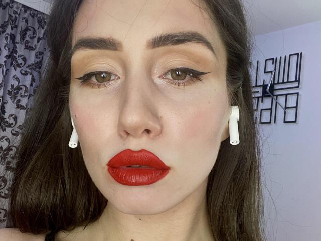 MissScarlettXo has red lipstick, white earbuds, brown eyes with bold eyeliner, and a serious expression. Her brown hair is down, and she has a light skin tone.