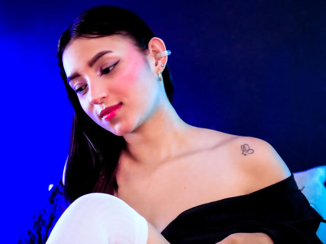 MaraLewis with dark hair, pink blush, black off-shoulder top, small heart tattoo on shoulder, holding white balloon, blue and pink lighting, slight smile, red lipstick, ear piercing.