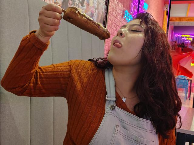 Noemi Thornee, avec ses cheveux bruns bouclés, son pull orange en maille côtelée et sa combinaison blanche, lèche un long hot-dog croûté qu'elle porte à sa bouche. Sa langue est dehors, les yeux fermés..