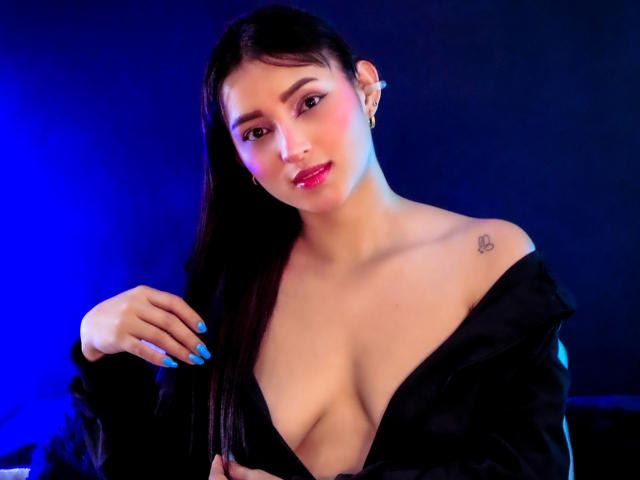 MaraLewis with long black hair, blue nails, black off-shoulder top, revealing cleavage, small heart tattoo, blue and purple lighting, smiling, pink lips, earrings.