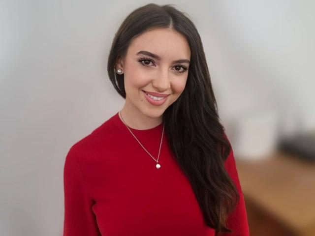 HarperSexy-hot with long dark hair, red dress, silver necklace, and pearl pendant, smiling softly at the camera. Boobs are not visible. She's cute and has a pretty face.