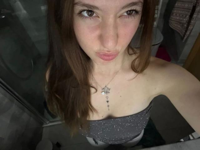 HarperSexy-hot has pale skin, brown eyes, and pouty lips. She's wearing a strapless gray top with sparkles, showing her small tits, and has a star necklace.