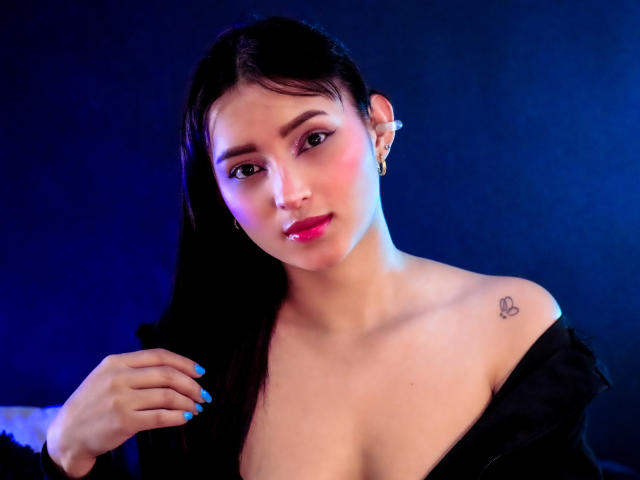 MaraLewis with black hair, blue nails, pink lipstick, gold earrings, heart tattoo on right shoulder, black off-shoulder top, looking at camera, blue light, soft smile, slight blush.