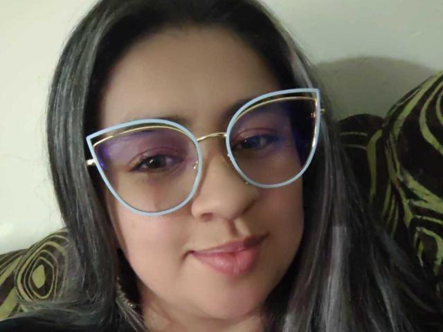 MelindaBoobs with medium skin, dark straight hair, purple eyeshadow, and blue-rimmed glasses, smiling slightly, showing subtle makeup, and a relaxed expression.