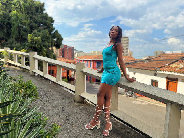 SabrinaDevil, dark-skinned, wearing a tight, turquoise, mini-dress, and white, strappy, high-heeled sandals, leans on a white railing, with green plants on the left.
