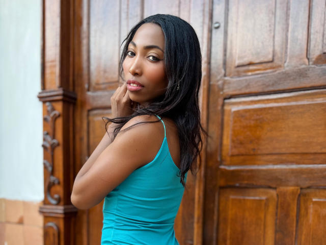 SabrinaDevil with dark skin, long black hair, wearing a tight teal dress, looking over her shoulder, touching her chin, wooden door in background. Her tits are slightly visible.