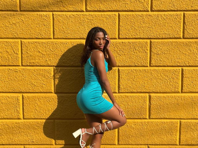 SabrinaDevil in a tight, teal dress, standing against a yellow brick wall, showing her ass, with white, lace-up heels, and her hand in her long, dark hair.