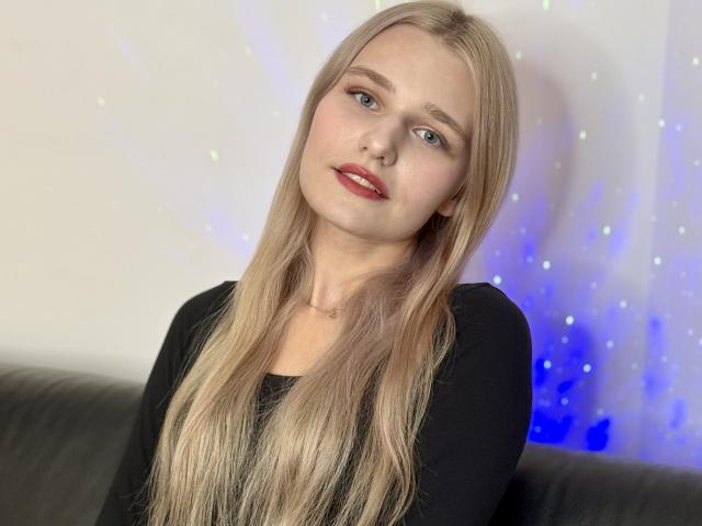 RebeccaChamb has long blonde hair, blue eyes, and red lipstick. She's wearing a black top, sitting on a black couch with a subtle blue and white light effect.