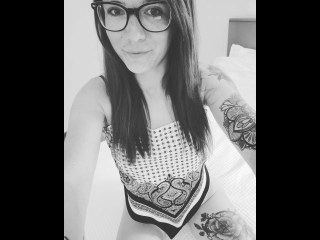 Solveig-hot with glasses, long dark hair, wearing a patterned top, showing tattoos on her arm and thigh, smiling at the camera in a black-and-white selfie.