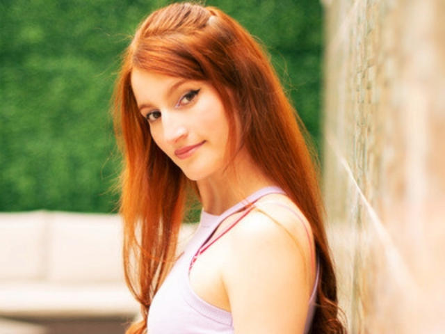 RitaJonness with long red hair, light skin, wearing a purple tank top, standing against a wall, looking at the camera with a slight smile, showing her tits.