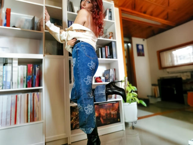 ZoeeMar con cabello rojo, blusa blanca con un hombro descubierto, jeans azules con estampado floral, botas negras, de pie sobre una pierna, apoyada contra una estantería blanca, trasero atractivo, de espaldas a la cámara, en una habitación acogedora..