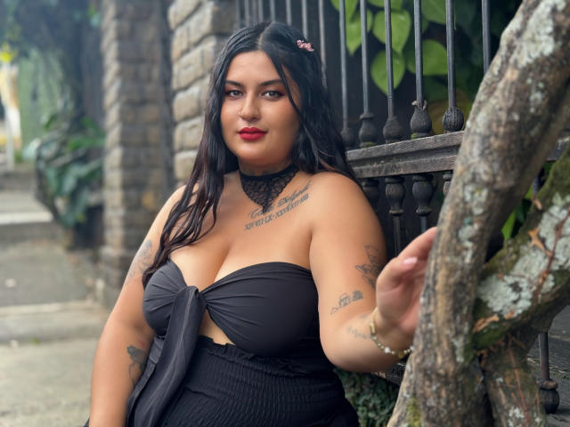 LilyRocks with big boobs, black strapless dress, tattoos, red lipstick, dark hair, choker, leaning on iron fence, right arm raised, tree in foreground, soft light.