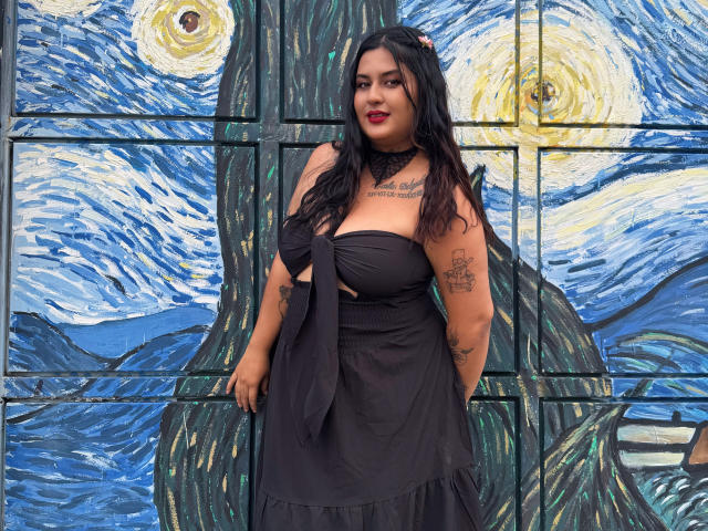 LilyRocks has black hair, red lipstick, and tattoos, wearing a strapless black dress with a bow, standing in front of a blue and yellow painted wall. Boobs visible.