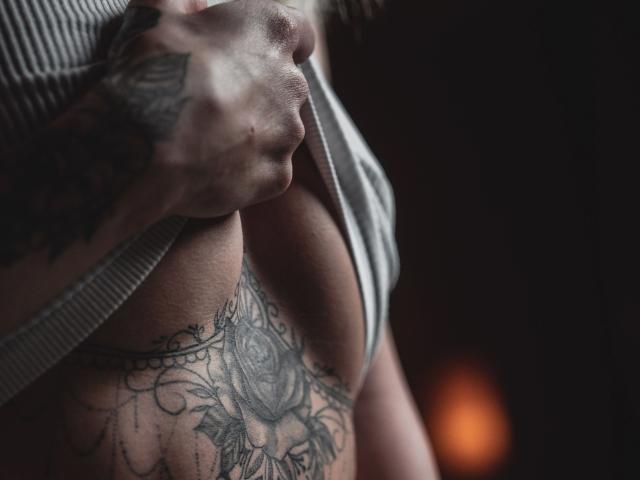 Younglily-hot's hand pulls up a striped shirt, revealing a large rose tattoo covering her tits and a detailed chest piece. Her skin is light, tattoos are dark.