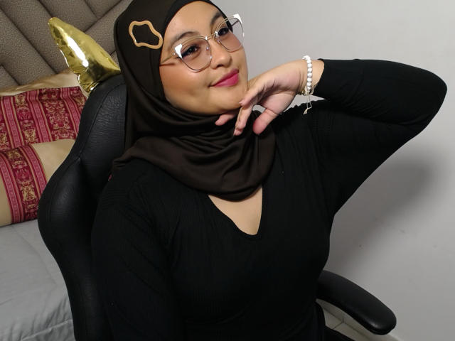 HelueJamil has brown skin, wearing glasses, a black hijab with a gold ornament, black long-sleeve top, and a pearl bracelet, sitting in a black chair, smiling.