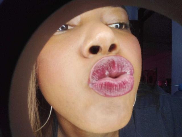 MacarenaBoobs with dark brown skin, pouty pinkish-purple lips, slightly upturned nose, blue eyes, and hoop earrings, wearing a black hat, making a kissing face.