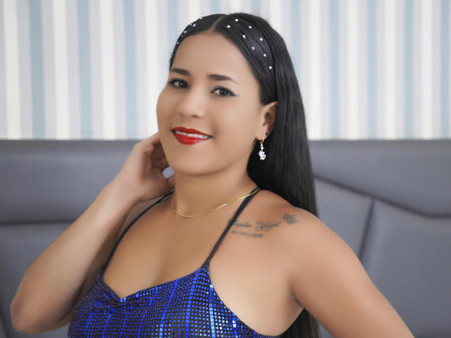 AlisLambert has dark hair, red lipstick, and a blue sequined top. She's smiling, with a tattoo on her right shoulder and a gold necklace. Her tits are small.