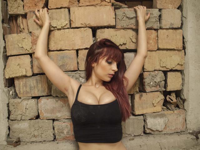 A mão de SensualCrissa, com a pulseira de contas vermelhas, toca suas calcinhas de renda preta, revelando suas coxas lisas e claras. Seus dedos puxam levemente a renda, mostrando sua pele macia..