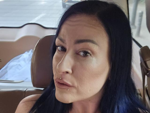 GennyRock ha i capelli blu scuro, gli occhi marroni e un piercing al naso. Ha un piercing al labbro e sembra leggermente confusa. È seduta in un'auto con sedili in pelle marrone..