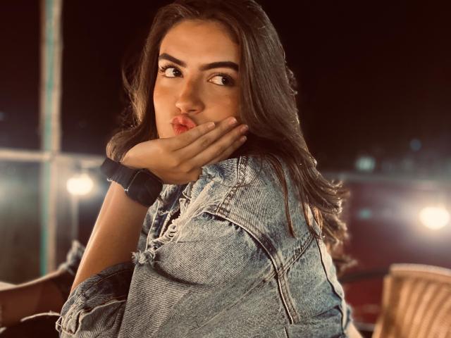 Nicolexa with long dark hair, wearing a denim jacket, pouting with her hand on her cheek, brown eyes looking sideways, wearing a black smartwatch, in a dimly lit setting.