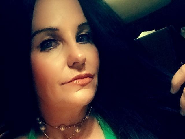 Wickedcrystal has dark hair, light skin, and a subtle smile. She's wearing a green top and a gold necklace. Her eyebrows are dark and she's looking slightly to the side.