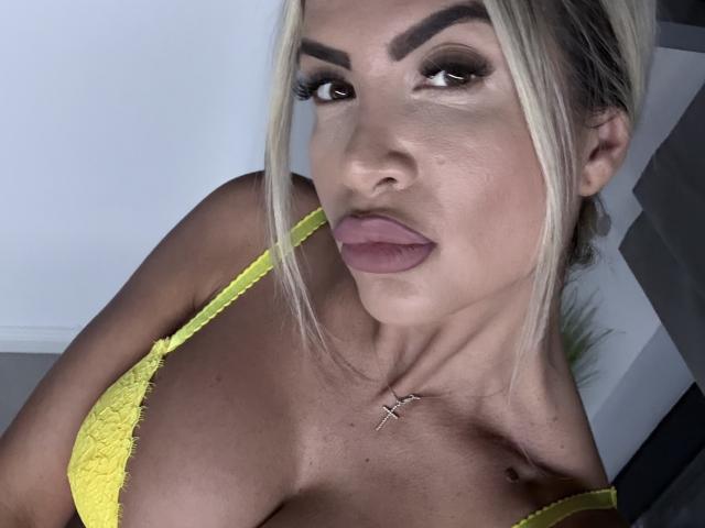 Lellie with blonde hair, thick eyebrows, and pink lips, wearing a yellow lace bra, showing her big boobs, a cross necklace, and a small tattoo below her right breast.