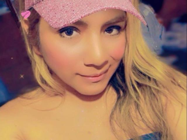 ChelseaForTranny with blonde hair, pink glitter headband, and a cute smile, showing her soft, pink-tinted tits in the selfie. She has sparkles on her skin.
