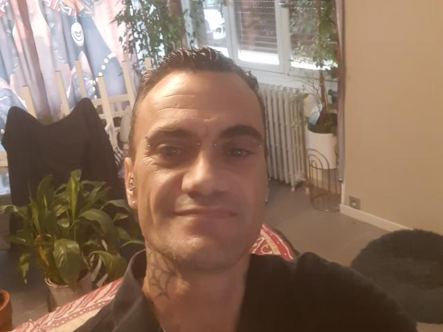 KevinLatrique, short dark hair, slight smile, tattoo on neck, black shirt, small ear piercings, medium skin, white wall, window, plant, radiator, black chair, and curtain in background.