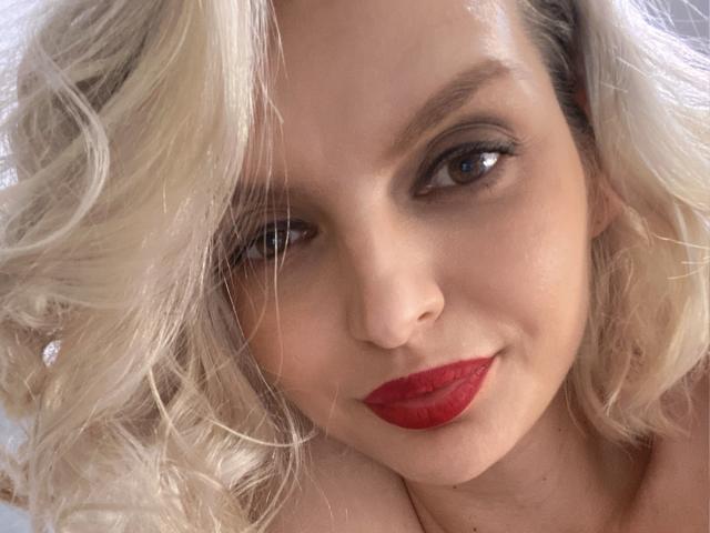 DouceAmelia with blonde curly hair, red lipstick, and brown eyes, smiling softly. Her fair skin and subtle makeup highlight her pretty face. She's looking directly at the camera.