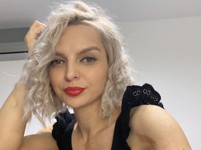 DouceAmelia with curly white hair, red lipstick, black top, looking at camera, right hand in hair, white background, slightly smiling, light skin, subtle makeup, medium breasts.