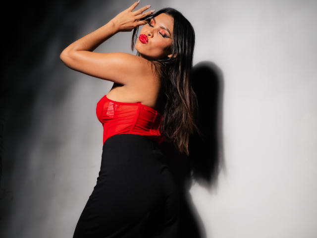 MarceBetancur with long brown hair, red sheer top, black skirt, raised arm, and red lipstick, posing against a gray wall, showing her bare ass and sexy face.