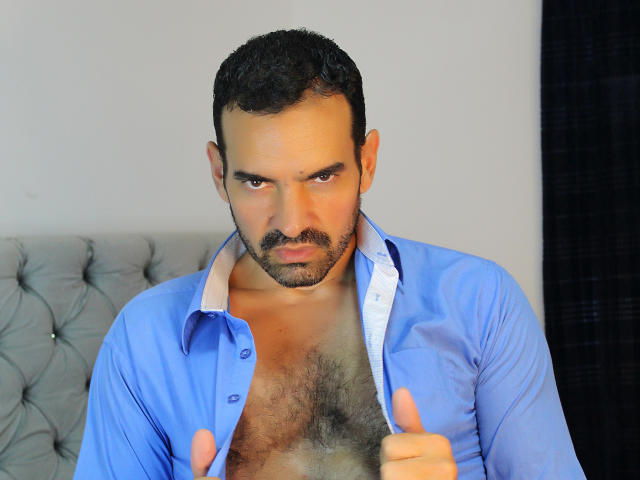 XRawForU with a serious expression, wearing an unbuttoned blue shirt, showing a hairy chest, and pointing at the camera with both hands. Dark curly hair, stubble.