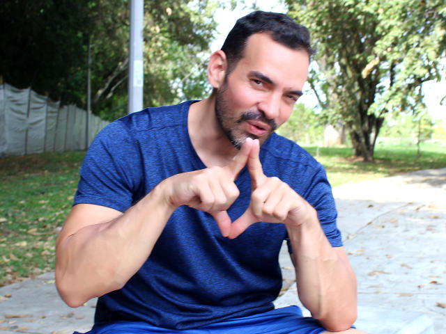 XRawForU, a muscular guy with a dark beard, wearing a blue shirt, sits making a heart shape with his fingers. He's slightly smiling, with short, dark hair.