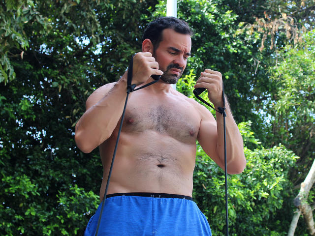 Muscular XRawForU with dark hair and beard, shirtless, holding black jump rope, wearing blue shorts, standing in green outdoor setting, looking focused, showing hairy chest and abs.