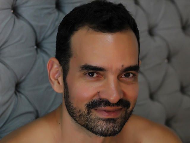 XRawForU has a chubby face, dark curly hair, a small mustache, and a beard, with a slight smile, and a small mole above his right eyebrow. Shirtless.