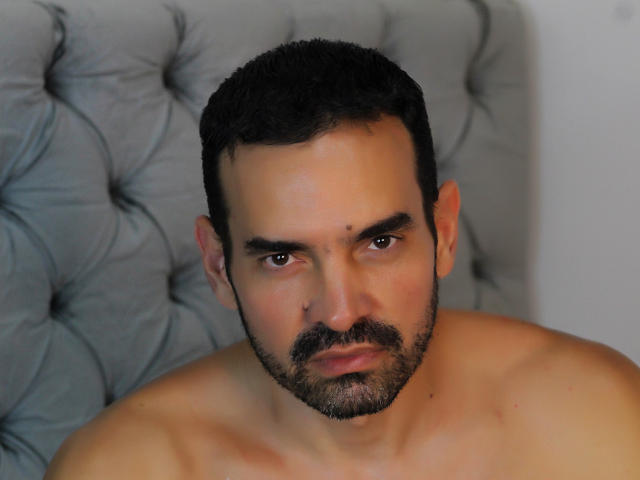 XRawForU has a serious expression, dark short hair, and a beard, with a shirtless, muscular upper body, and a tufted grey headboard in the background.