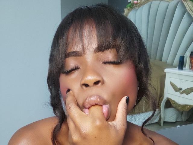 O rosto marrom escuro de LindaBird, com olhos fechados, blush rosa e esmalte preto, sugando o dedo com a mão perto da boca, em um quarto..