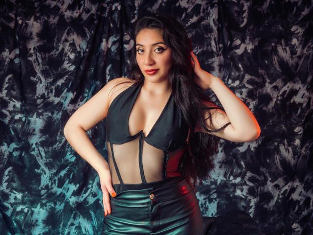 ArianaZahar with long dark hair, wearing a black halter top with mesh and black leather pants, hand in hair, red lipstick, confident smile, medium tits, dark patterned background.