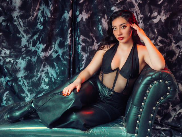 ArianaZahar with big tits, wearing a black, cut-out halter top and black leather pants, lounging on a green, studded leather couch, touching her dark hair.