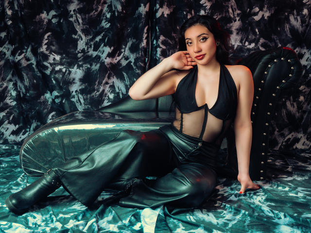 ArianaZahar with dark hair, wearing a black halter top, exposing her boobs, and black leather pants, sitting on a green, shiny, inflatable lounge, touching her chin.