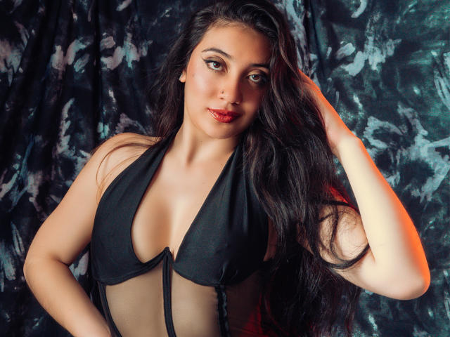 ArianaZahar with long black hair, wearing a black halter top, showing her tits, red lipstick, and a confident expression, hand in hair, against a dark, textured background.
