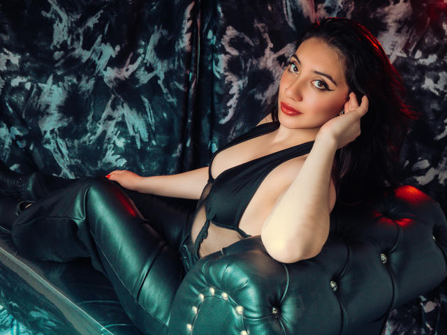 ArianaZahar with pale skin, dark hair, and red lipstick, lounging on a green tufted leather couch in a black halter top, exposing her tits. She's touching her head.
