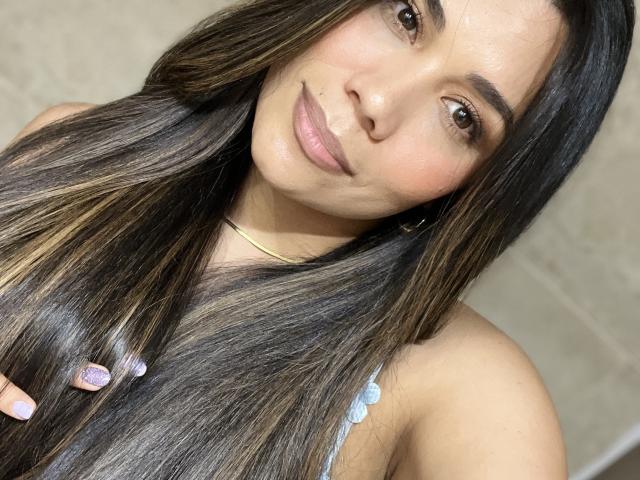 Sheyleeya with long dark brown hair, slightly glossy, holding it on the left. She has brown eyes, light skin, and a subtle smile with pink lipstick. Her tits are not visible.