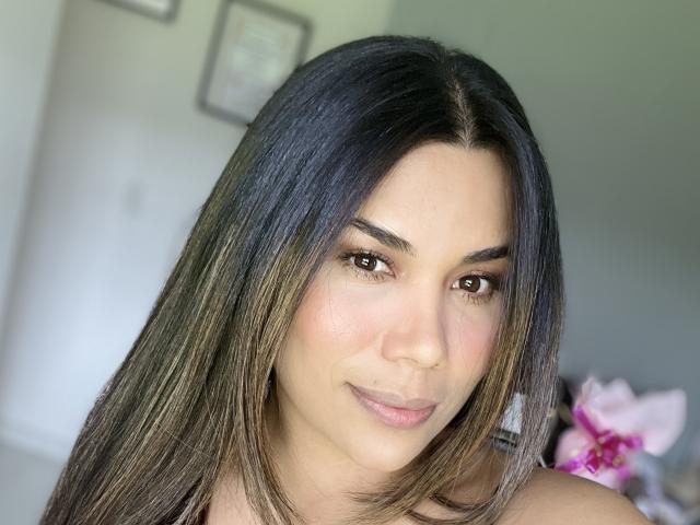 Sheyleeya has long, dark brown hair with subtle highlights, brown eyes, and a slight smile, showing her cute, round nose and soft pink lips. Her boobs are partially visible.